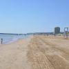 Spiaggia sabbiosa con mare calmo, alcune persone in lontananza e un edificio alto sullo sfondo.