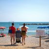 Famiglia con passeggino cammina verso una spiaggia con ombrelloni colorati e mare sullo sfondo.