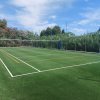 Campo da pallavolo all'aperto con erba finta e rete, circondato da alberi e cielo sereno.