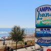 Un cartello di un campeggio vicino a una spiaggia. Persone camminano lungo la strada.