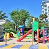 Parco giochi con scivoli colorati su sabbia, palme e un edificio sullo sfondo.