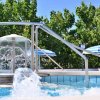 Una piscina con scivolo, giochi d’acqua e ombrelloni a righe blu, circondata da alberi verdi.