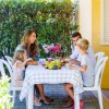 Famiglia a tavola all'aperto con frutta e fiori, davanti a una siepe verde.