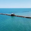 Lungo molo di legno che si estende nel mare, affiancato da una spiaggia con ombrelloni.