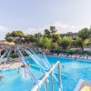 Piscina all'aperto con giochi d'acqua e persone che nuotano, circondata da alberi e sedie a sdraio.