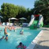 Una piscina gremita di persone con uno scivolo gonfiabile verde e sedie sdraio sotto ombrelloni.
