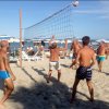 Persone giocano a pallavolo su una spiaggia soleggiata con ombrelloni e mare sullo sfondo.