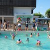 Piscina con bambini che nuotano e adulti sul bordo. Una persona in costume da mascotte sullo sfondo.