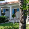 Bungalow bianco con finestre blu, veranda e sedie su un prato verde.