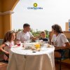 Famiglia di quattro persone che fa colazione su un terrazzo soleggiato.