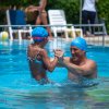 Uomo e bambina con cuffie blu giocano sorridenti in una piscina.