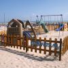 Area giochi per bambini su spiaggia sabbiosa, con altalene e scivoli circondati da una staccionata.