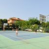 Due persone giocano a tennis su un campo all'aperto, circondato da alberi e edifici.