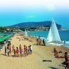Spiaggia affollata con vele bianche e verdi, montagne sullo sfondo.