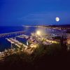 Vista notturna di un porto illuminato con una luna piena e mare calmo.