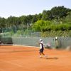 Due persone giocano a tennis su un campo in terra battuta, circondato da alberi.