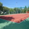 Campo sportivo all'aperto con superficie rossa, reti di protezione e alberi intorno.