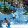 Persone si divertono in piscina con scivolo e spruzzi d'acqua.