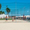 Persone giocano a beach volley su sabbia con sfondo di ombrelloni e palma.