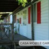 Casa mobile bianca con persiane rosse e veranda in legno, circondata da alberi.