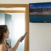 Una donna guarda un televisore a parete con vista aerea di una spiaggia, telecomando in mano.