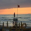 Tramonto sul mare con una bandiera rossa e ombrelloni chiusi in spiaggia.