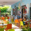 Una terrazza con tavoli e sedie colorate. Persone sedute e vegetazione visibile sullo sfondo.