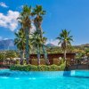 Piscina con fontane, palme e un ponte di legno, sfondo montuoso sotto un cielo azzurro.