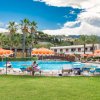 Piscina all'aperto con ombrelloni arancioni e lettini, circondata da palme e colline verdi.