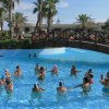 Persone svolgono esercizi in piscina, ponte di legno e palme.