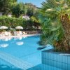 Lato di una piscina con piante rigogliose e lettini con ombrelloni sullo sfondo.
