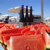 Fette di anguria e bottiglie di vino su un tavolo sotto ombrelloni da spiaggia.