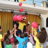 Bambini giocano con palloncini rossi insieme a una mascotte in un'area con tende gialle.