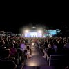 Platea notturna con pubblico seduto e palco illuminato sullo sfondo.