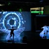 Due performer danzano sul palco con costumi a LED, sfondo di proiezioni luminose.