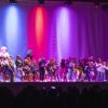 Gruppo numeroso di bambini su un palco colorato durante una performance teatrale o musicale.