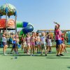 Bambini e animatori danzano allegri in un parco giochi all'aperto con scivoli colorati.