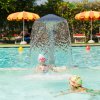Due bambine giocano in piscina sotto una fontana d'acqua. Ombrelloni sullo sfondo.