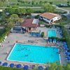 Piscina con persone e ombrelloni blu, circondata da alberi e edifici.