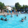 Persone giocano in una piscina sotto un cielo sereno. Palme e sdraio sullo sfondo.