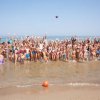 Un grande gruppo di persone salta gioiosamente in riva al mare con le mani alzate.