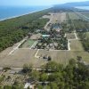 Veduta aerea di un villaggio turistico con piscine, campi sportivi e foresta lungo la costa.