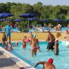 Gruppo di persone che giocano in piscina, con ombrelloni blu sullo sfondo.
