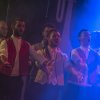 Gruppo di cantanti maschili sul palco con luci blu, vestiti formali.