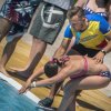 Istruttore aiuta una bambina a tuffarsi in piscina, con altre persone sullo sfondo.